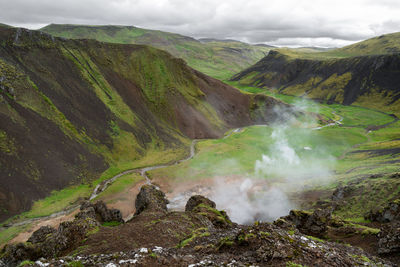 Steam smoke rises from boiling mud in a brown and green valley of geysir geothermal area, iceland.