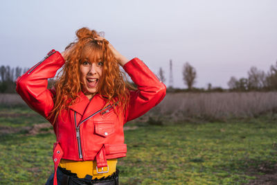 Young caucasian woman dressed in red leather windbreaker with a happy face screaming in the field.