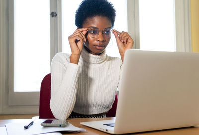 Concentrated young african american female in casual wear and eyeglasses looking at laptop screen and reading professional information while working at table in office