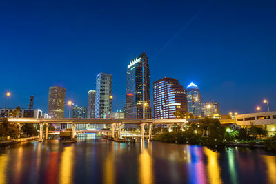 Illuminated buildings by river against blue sky at night