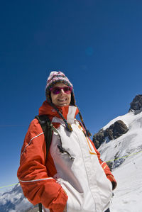 Low angle view of woman smiling at camera while standing on mountain against clear blue sky during winter