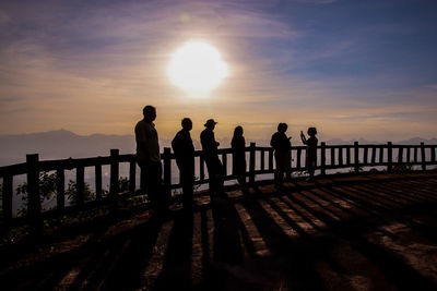 Silhouette people standing on railing against sky during sunset