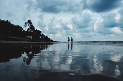 Rear view of friends standing at beach against cloudy sky