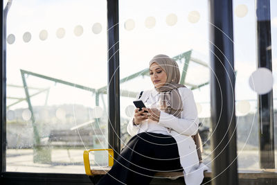 Woman in headscarf waiting at bus stop and using phone