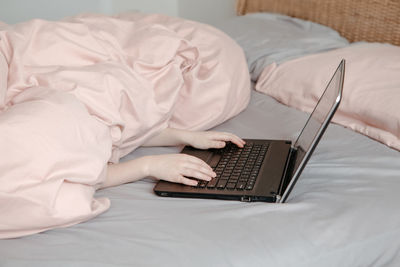 Bored funny child lying in bed under blanket and learning in virtual online school class. hands 