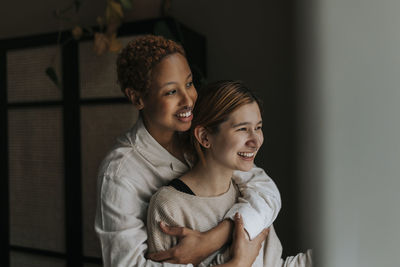 Smiling non-binary person embracing happy friend at home