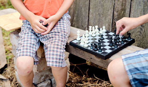 Two village boys play chess on the street, sitting on a bench, against the backdrop of an old barn