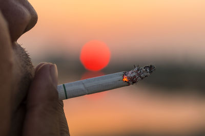 Close-up of hand holding cigarette against sky during sunset