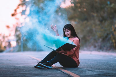 Smiling woman with blue smoke emitting book sitting on road