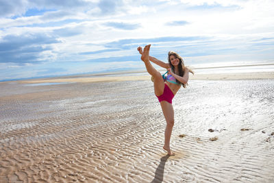 Full length portrait of young woman jumping at beach against sky