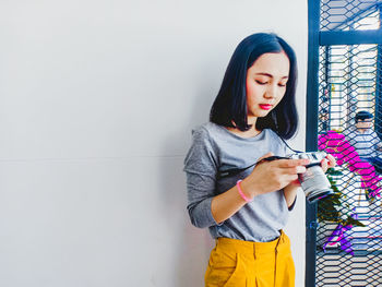 Young woman holding camera while standing against wall