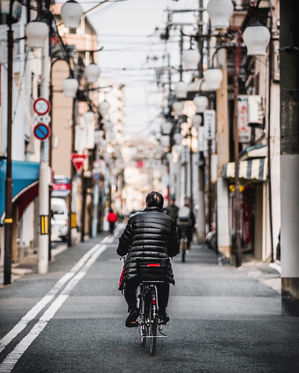 transportation, city, rear view, architecture, real people, the way forward, street, mode of transportation, building exterior, direction, bicycle, one person, lifestyles, land vehicle, built structure, road, focus on foreground, city life, sign, riding, outdoors