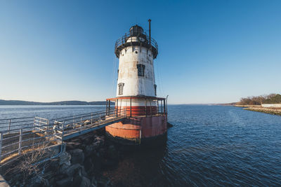 Lighthouse by sea against clear blue sky