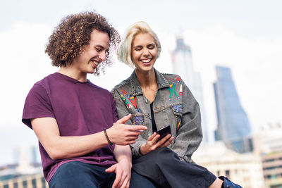 Smiling couple using phone while sitting against buildings in city