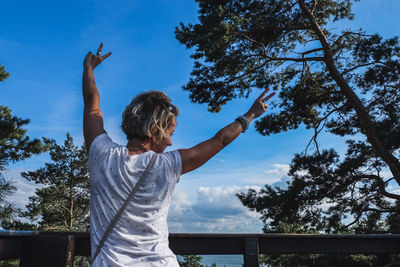 Rear view of woman with arms raised gesturing peace sign against trees and sky