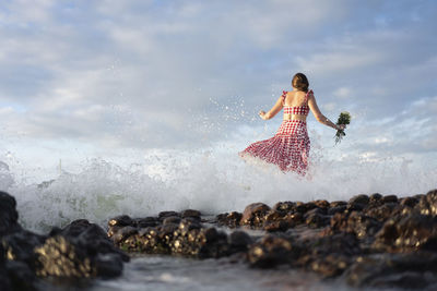 Rear view of woman standing on rock by sea against sky