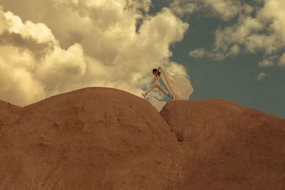 Low angle view of young woman jumping on mountain against cloudy sky