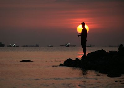 Silhouette man standing on rock by sea against sky during sunset