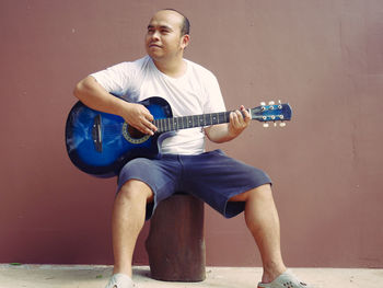 Full length of man playing guitar while sitting on log against wall