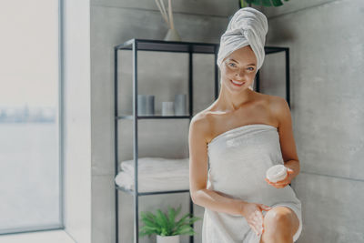 Portrait of smiling woman applying moisturizer while standing in bathroom