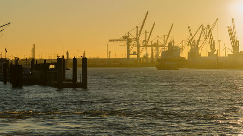 Silhouette cranes at harbor against sky during sunset