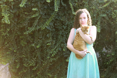 Portrait of young woman holding cat while standing against plants in park