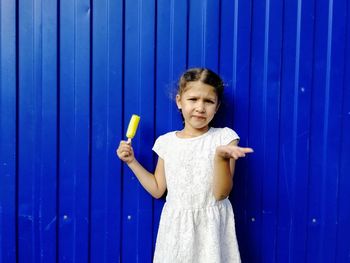 Portrait of cute girl holding flavored ice while gesturing against blue corrugated iron