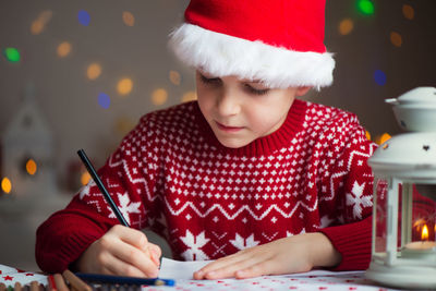 Close-up of cute boy wearing santa hat writing while sitting by oil lamp at home