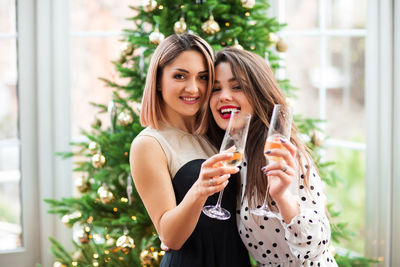Portrait of smiling young women holding champagne against christmas tree