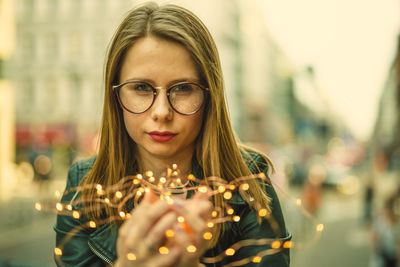 Close-up portrait of young woman holding illuminated string lights in city