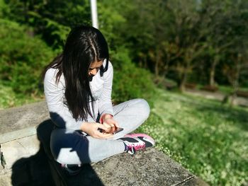 Woman using phone while sitting on resting wall against trees