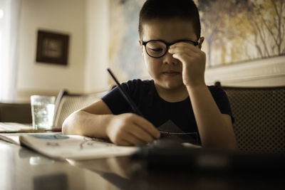 Boy with glasses draws in album with colored pencils, eye fatigue. glass of clean water on the table