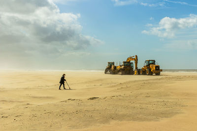 Woman walking against earth movers at beach against sky