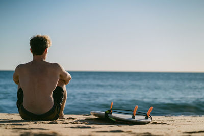 Rear view of shirtless man sitting on beach against clear sky