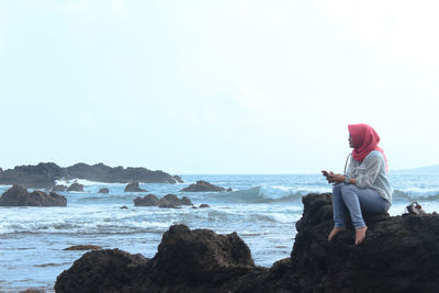 Woman sitting on rock at beach against sky