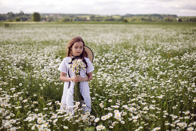 Portrait girl child in a white dress stands on a camomile field in a hat and bouquet of flowers