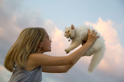 Young woman lifting cat against sky