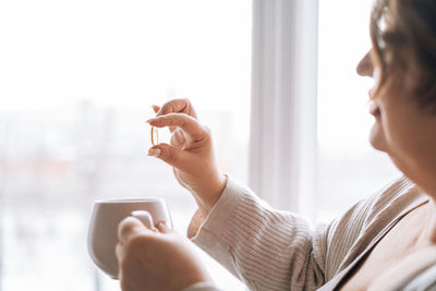 Close up photo of middle aged woman holding omega 3 capsule and mug of water in hands near window