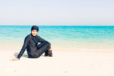Portrait of smiling young woman sitting on beach against clear sky