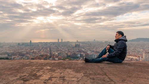 Side view of man sitting on city against sky during sunset