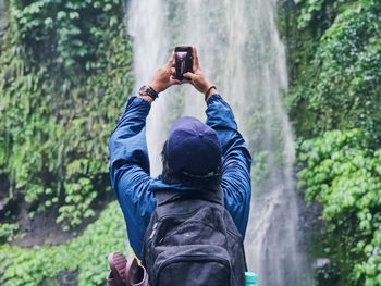 Rear view of man photographing waterfall through mobile phone
