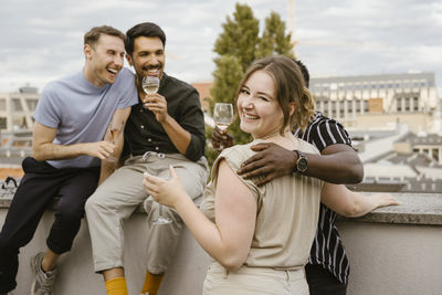 Portrait of smiling woman looking over shoulder while enjoying drinks with male friends on rooftop deck