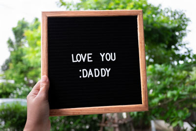 Cropped hand of person holding blackboard with text against trees