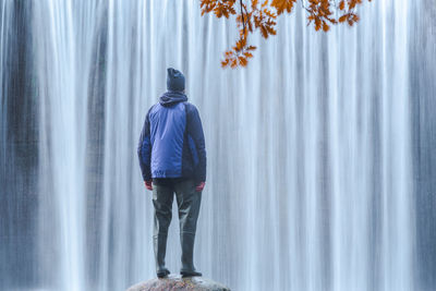 Back view of traveling male in warm clothes standing on boulder and admiring view of rapid waterfall in long exposure at lozoya river in guadarrama national park