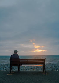 Rear view of man sitting on beach against sea during sunset