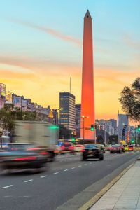 Cars on road by buildings against sky during sunset