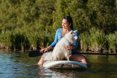 Happy young woman on the lake at early morning sitting on the sup board with her dog