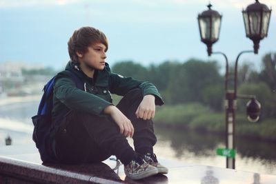 Thoughtful teenage boy sitting on retaining wall by street light against sky