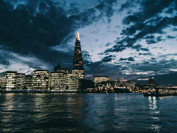 Illuminated buildings by river against cloudy sky