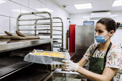 Young female baker positioning sheet on rack at bakery kitchen during pandemic